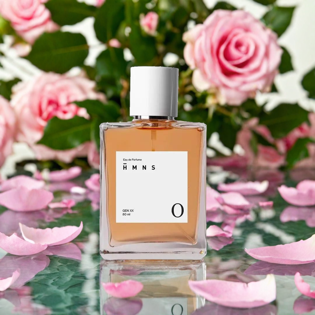 Botol parfum berada di antara kelopak bunga mawar pink dan bunga pink lainnya, dengan latar belakang kabur dari dedaunan hijau, menciptakan suasana segar dan romantis.