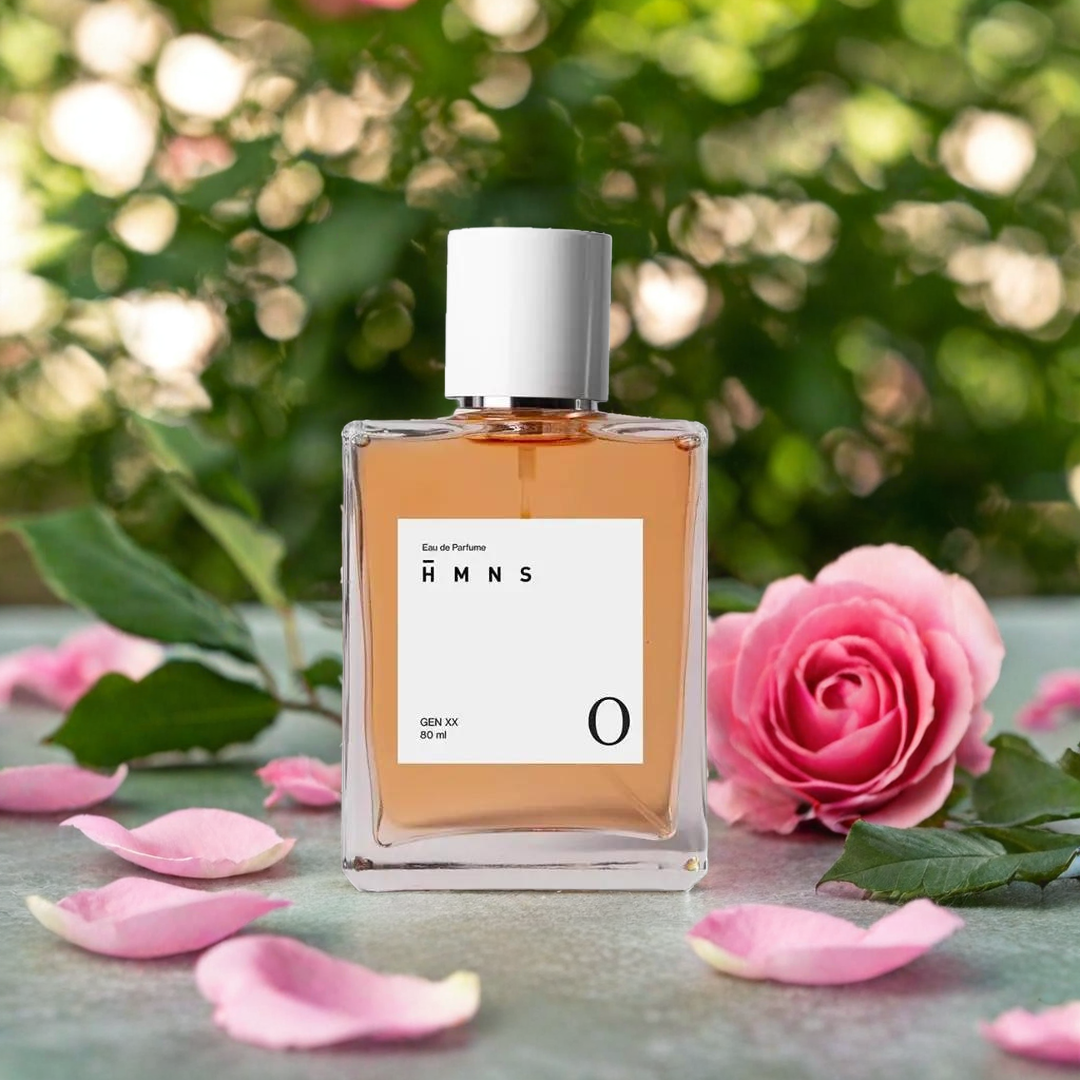 Botol parfum berada di antara kelopak bunga mawar pink dan bunga pink lainnya, dengan latar belakang kabur dari dedaunan hijau, menciptakan suasana segar dan romantis.